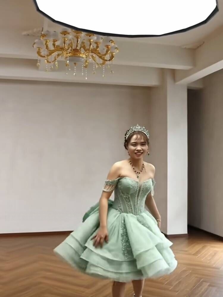 A smiling model wearing the completed mint green off-the-shoulder mini dress with a tiered ruffled skirt and a tiara, standing under a chandelier. A smiling model wearing the completed mint green off-the-shoulder mini dress with a tiered ruffled skirt and a tiara, standing under a chandelier.