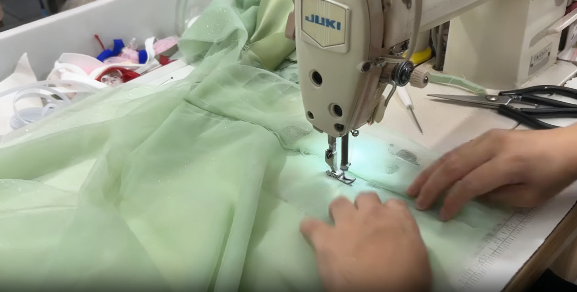 Close-up of a tailor sewing sheer mint green shimmer fabric with a JUKI sewing machine, hands guiding the material. Close-up of a tailor sewing sheer mint green shimmer fabric with a JUKI sewing machine, hands guiding the material.
