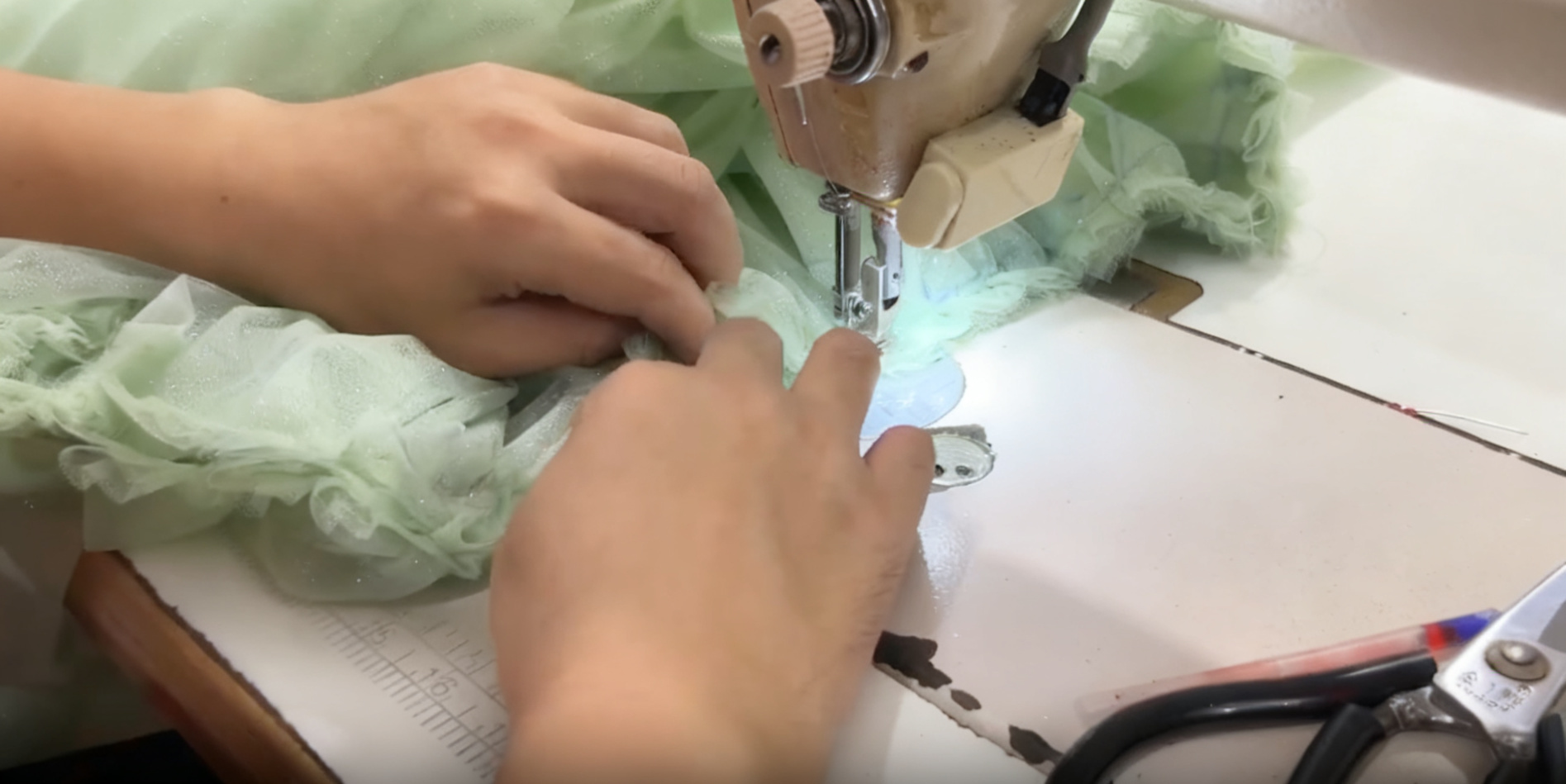 Hands feeding gathered ruffles of mint green tulle into an industrial sewing machine to create volume on the skirt. Hands feeding gathered ruffles of mint green tulle into an industrial sewing machine to create volume on the skirt.