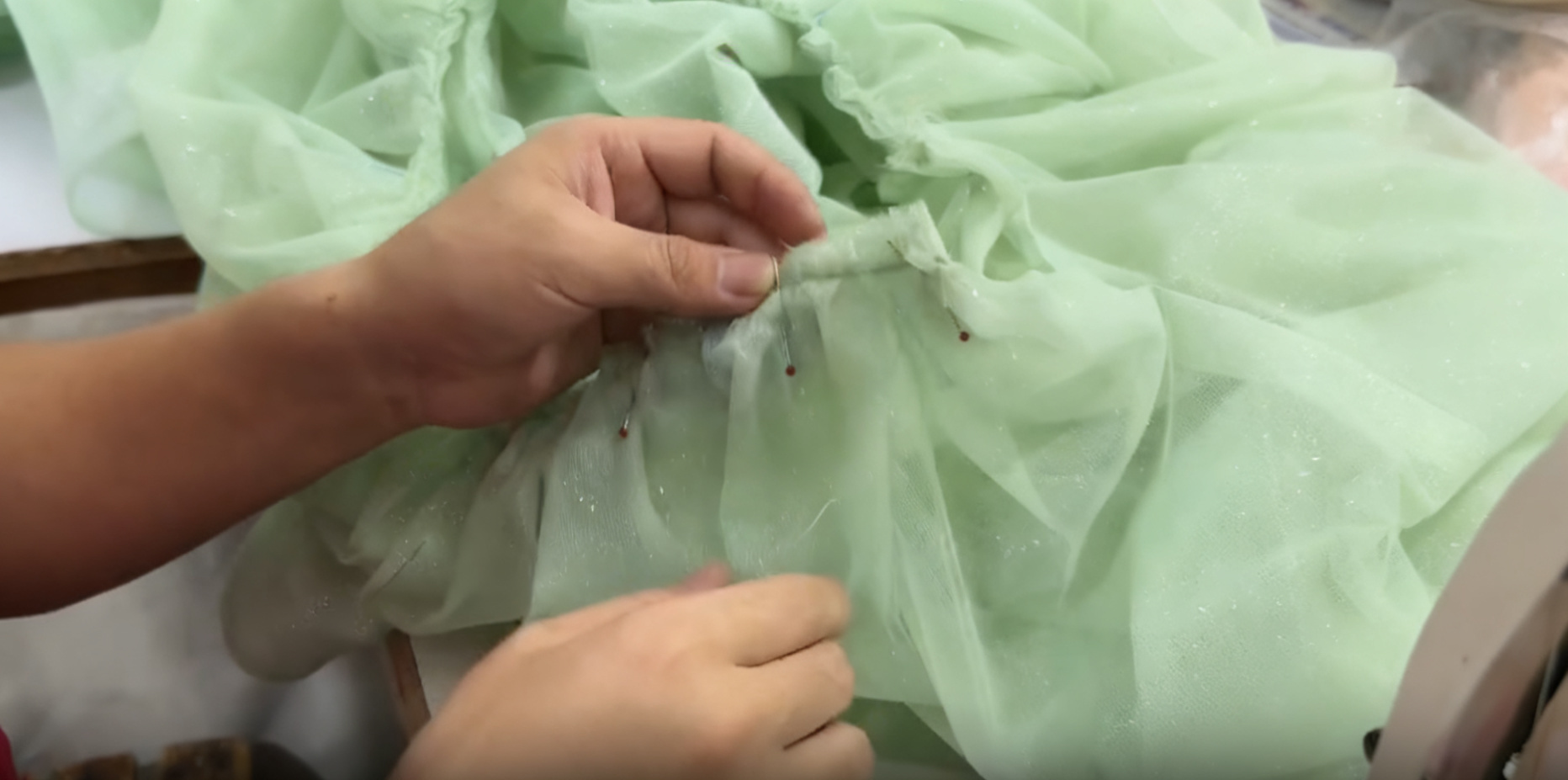 Close-up of a seamstress pinning folded pleats of mint green sheer fabric together with red-headed sewing pins. Close-up of a seamstress pinning folded pleats of mint green sheer fabric together with red-headed sewing pins.