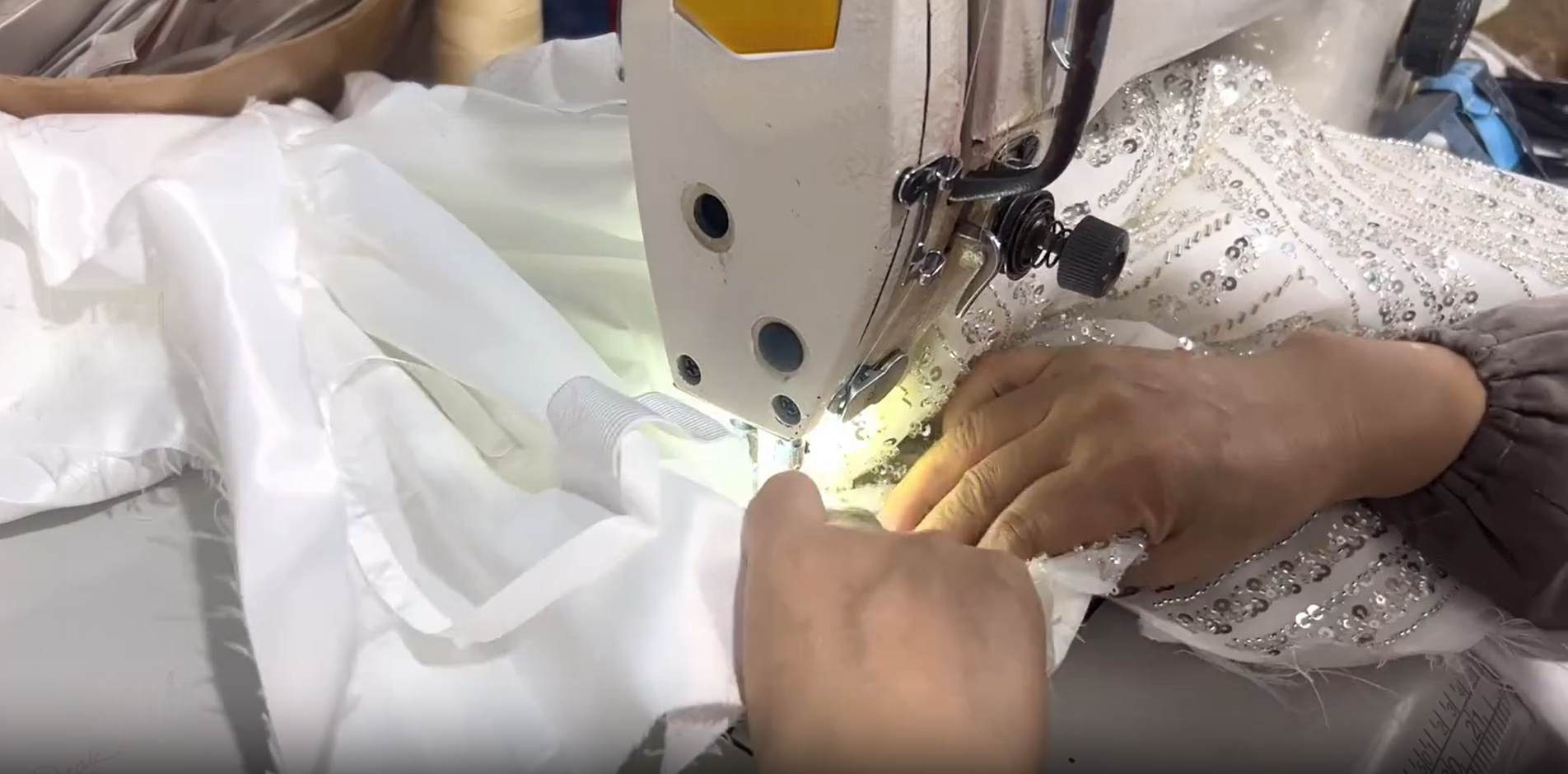 Hand guiding the structured, heavily embroidered white and silver bodice piece under the sewing machine needle to attach it to the inner lining layers. Hand guiding the structured, heavily embroidered white and silver bodice piece under the sewing machine needle to attach it to the inner lining layers.