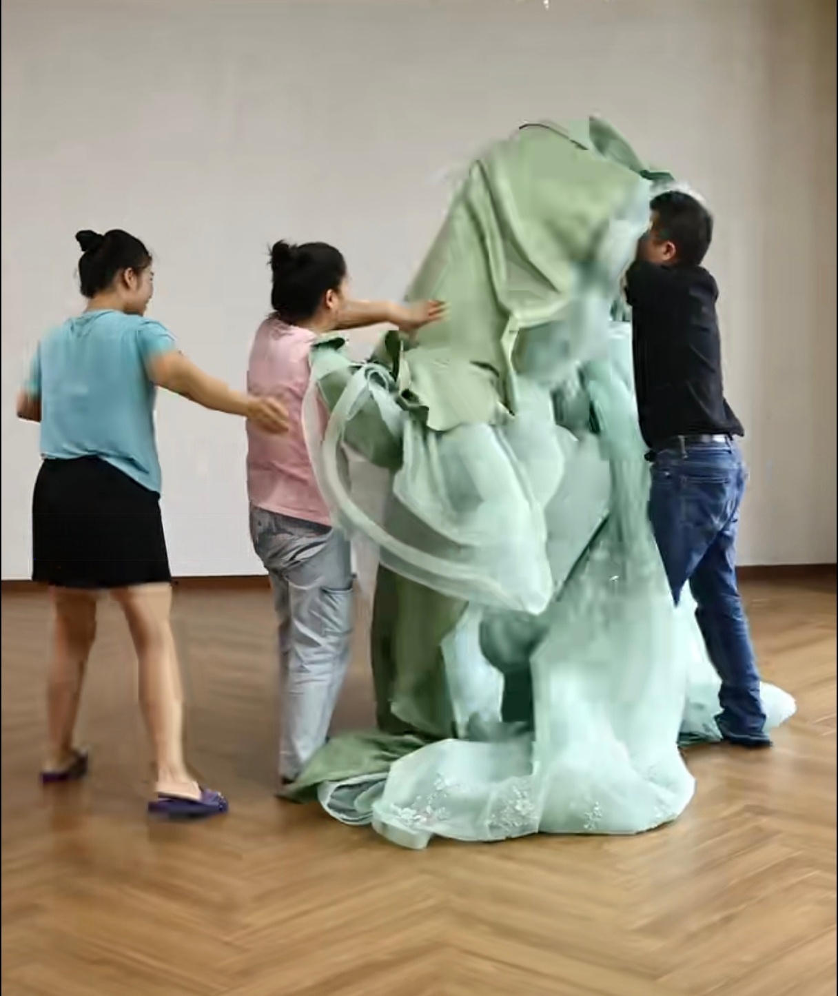 Three team members lifting the massive mint green dress to drape it over the dress form, working together to manage the huge volume of fabric. Three team members lifting the massive mint green dress to drape it over the dress form, working together to manage the huge volume of fabric.