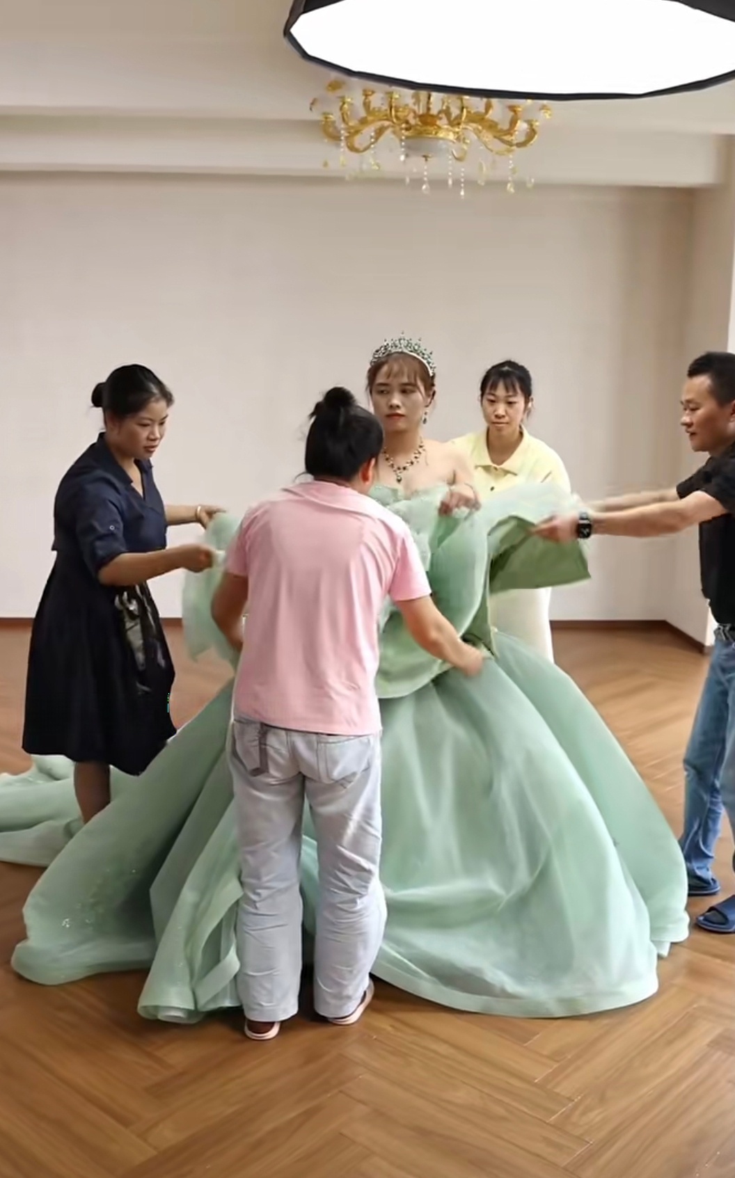 Four team members surrounding a model to help her put on the enormous floor-length mint green ball gown, managing the heavy layers of fabric. Four team members surrounding a model to help her put on the enormous floor-length mint green ball gown, managing the heavy layers of fabric.